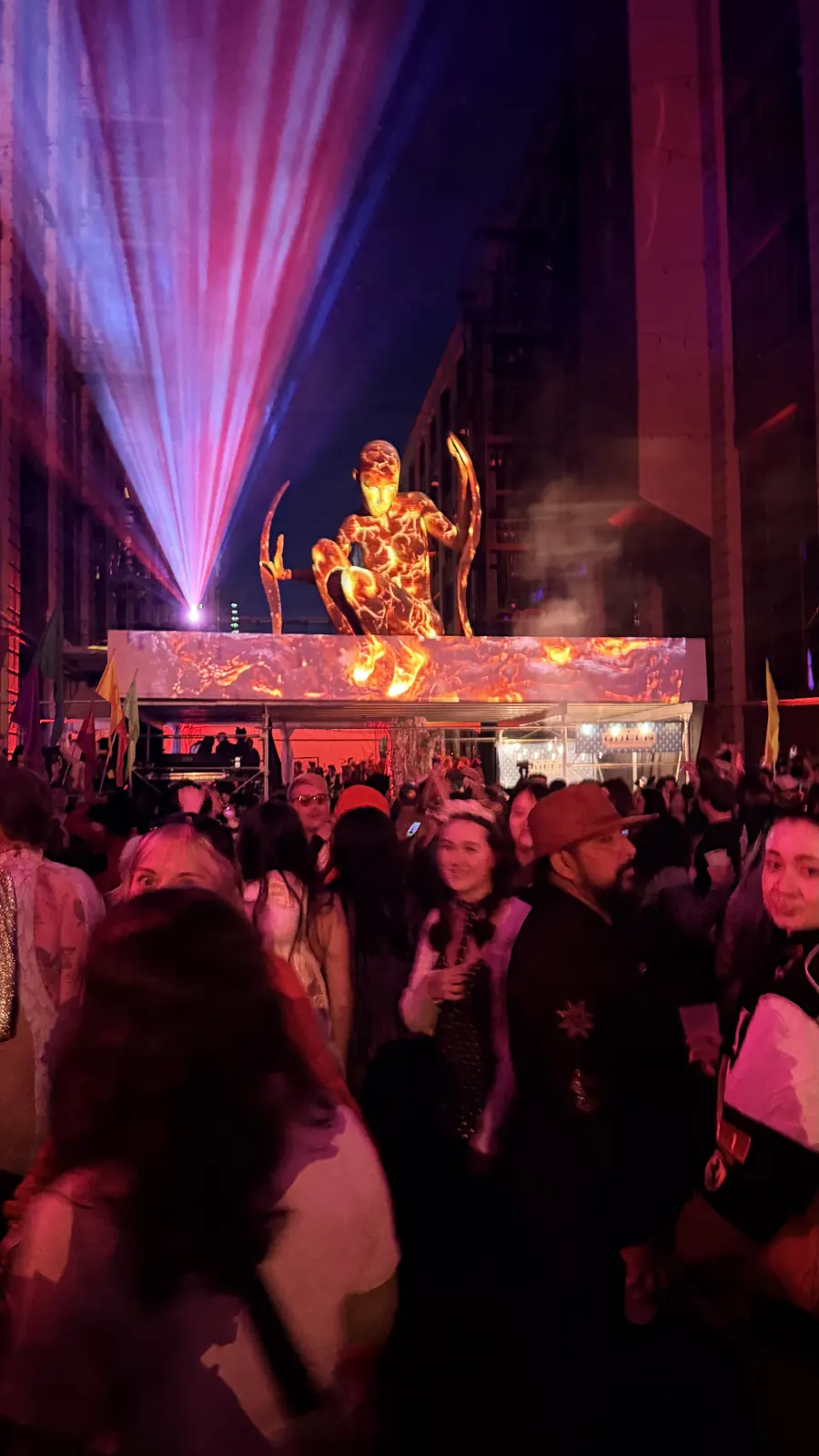 Colored beams illuminate a sculptural figure above the courtyard crowd.